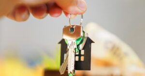 A hand holding keys with a house-shaped keychain over coins and a wallet