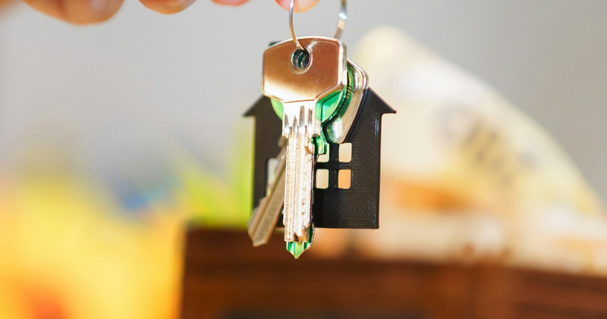 A hand holding keys with a house-shaped keychain over coins and a wallet